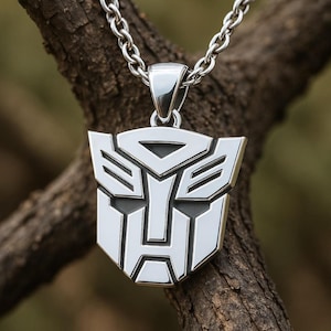 Könnte beinhalten: Ein silberfarbener Metallanhänger in Form des Autobot-Logos aus der Transformers-Reihe. Der Anhänger hat eine polierte, reflektierende Oberfläche mit vertieften, mattgrauen Details und hängt an einer passenden silberfarbenen Metallkette.