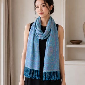 Pode incluir: Um cachecol azul-claro com um padrão paisley detalhado em tons de roxo e dourado, finalizado com franjas longas em ambas as extremidades. O cachecol está drapeado ao redor do pescoço sobre um vestido preto sem mangas, exibindo o design.