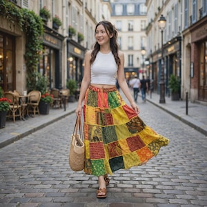 Puede incluir: Una camiseta blanca sin mangas combinada con una falda larga de patchwork hasta los tobillos, con una mezcla vibrante de cuadrados de tela amarillos, verdes y rojos con varios patrones. Un bolso de paja tejido y sandalias de cuero marrón.