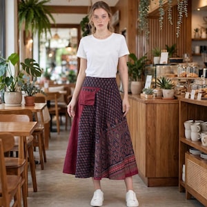 Puede incluir: Una camiseta blanca de manga corta con cuello redondo combinada con una falda midi oscura que presenta un panel lateral granate, un pequeño bolsillo de parche y un patrón geométrico repetitivo en tonos granate, azul y dorado, con zapatillas blancas.