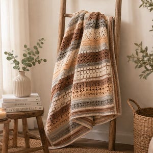 May include: A crocheted throw blanket featuring horizontal stripes in shades of brown, cream, tan, and grey. The blanket displays a variety of intricate stitch patterns, including openwork and solid rows, draped over a wooden ladder frame.