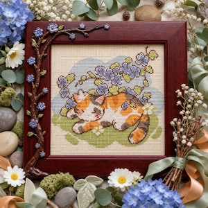 May include: A square wooden frame featuring a cross-stitch embroidery of a calico cat resting on green grass under blue flowers. The frame is decorated with a sculpted brown branch with small blue flowers along the top and left side.