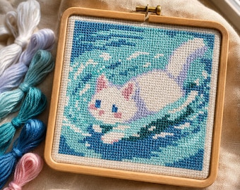 May include: A square wooden embroidery hoop containing a cross-stitch pattern of a white cat swimming in blue water. The cat has pink ears and blue eyes. Several skeins of embroidery floss in shades of white, blue, and pink are placed nearby.