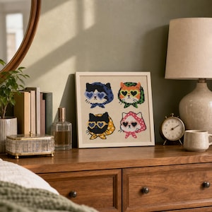 May include: A square framed cross-stitch embroidery featuring four pixel-art style cats wearing heart-shaped sunglasses and patterned headscarves. The cats are arranged in a grid, with colors including blue, orange, black, and pink on a light fabric.