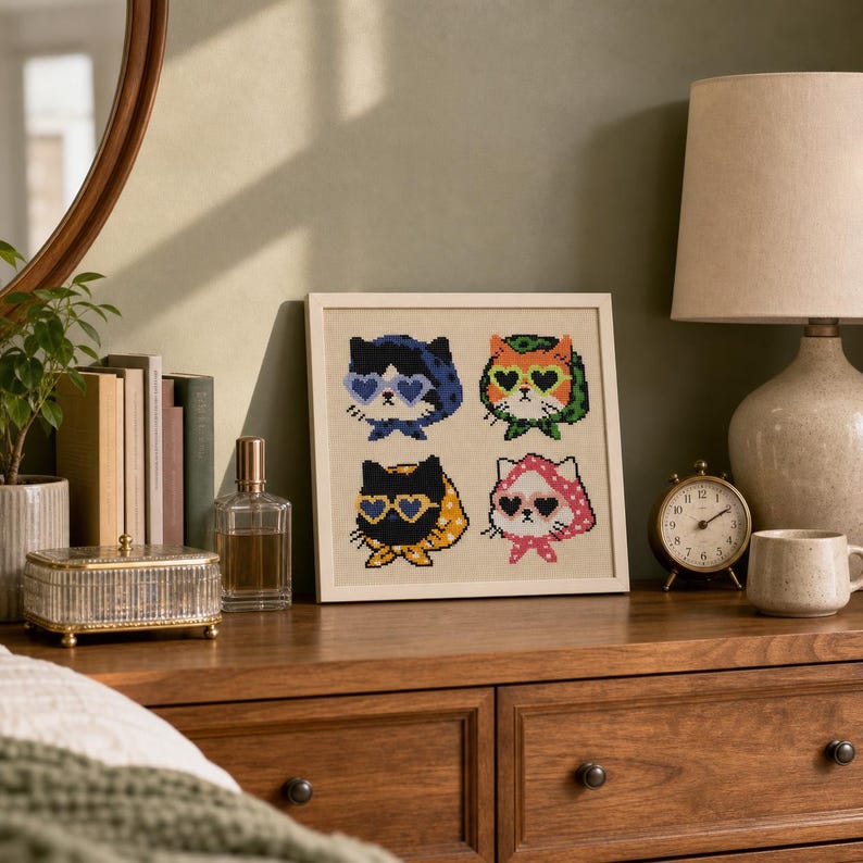 May include: A square framed cross-stitch embroidery featuring four pixel-art style cats wearing heart-shaped sunglasses and patterned headscarves. The cats are arranged in a grid, with colors including blue, orange, black, and pink on a light fabric.