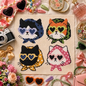 May include: A cross-stitch embroidery on cream fabric featuring four cat heads wearing headscarves and heart-shaped sunglasses. The cats are colored black with blue, orange with green, black with yellow, and white with pink, each in a distinct pattern.