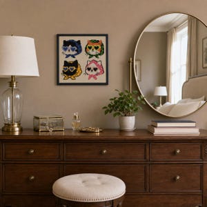 May include: A square framed cross-stitch art piece featuring four stylized cats wearing heart-shaped sunglasses and headscarves in blue, orange, yellow, and pink, displayed on a wall above a wooden dresser with a lamp and mirror.