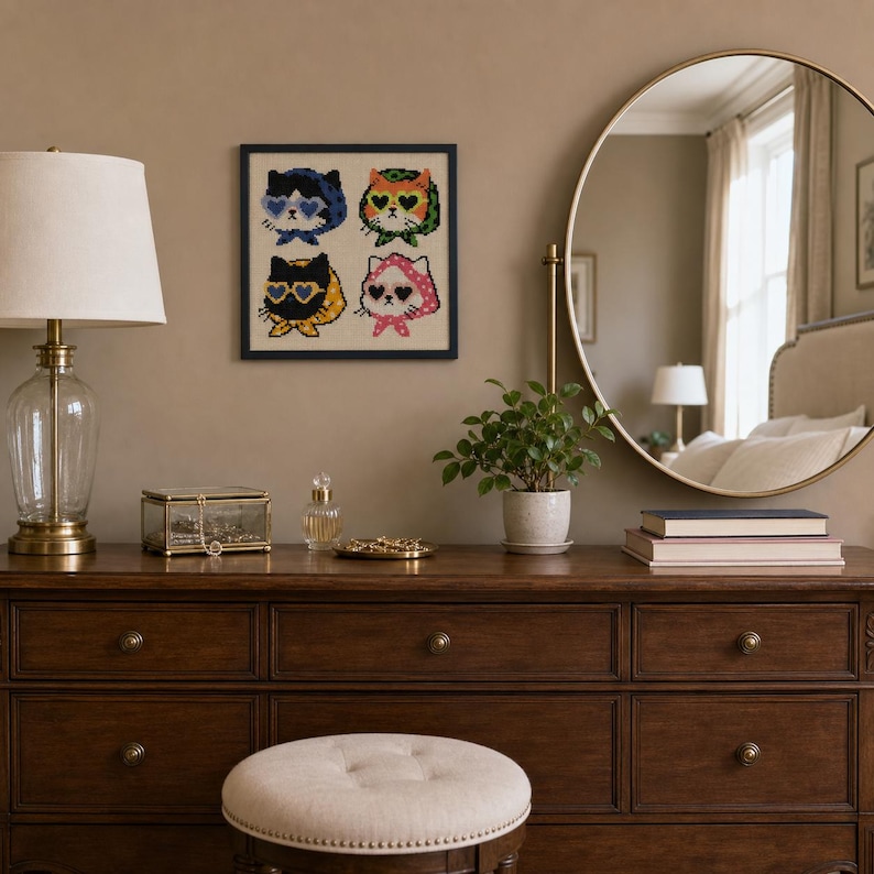 May include: A square framed cross-stitch art piece featuring four stylized cats wearing heart-shaped sunglasses and headscarves in blue, orange, yellow, and pink, displayed on a wall above a wooden dresser with a lamp and mirror.