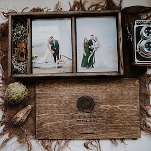 May include: A wooden box with a hinged lid, containing two framed photographs of a wedding couple and a group of people. The box also includes a USB drive and a vintage camera. The box is labeled "SYCAMORE FILM CO."
