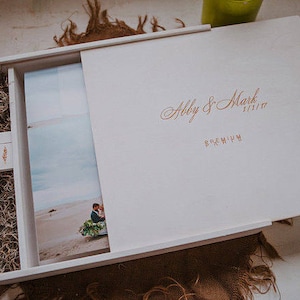 May include: White wooden box with a hinged lid. The lid is open and shows a photo album with a photo of a couple on the beach. The lid is inscribed with the text "Abby & Mark 11.11.17 PREMIUM".