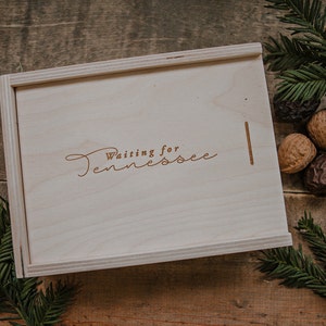 May include: A wooden box with the words "Waiting for Tennessee" engraved on the lid. The box is sitting on a wooden surface with pine branches and walnuts.