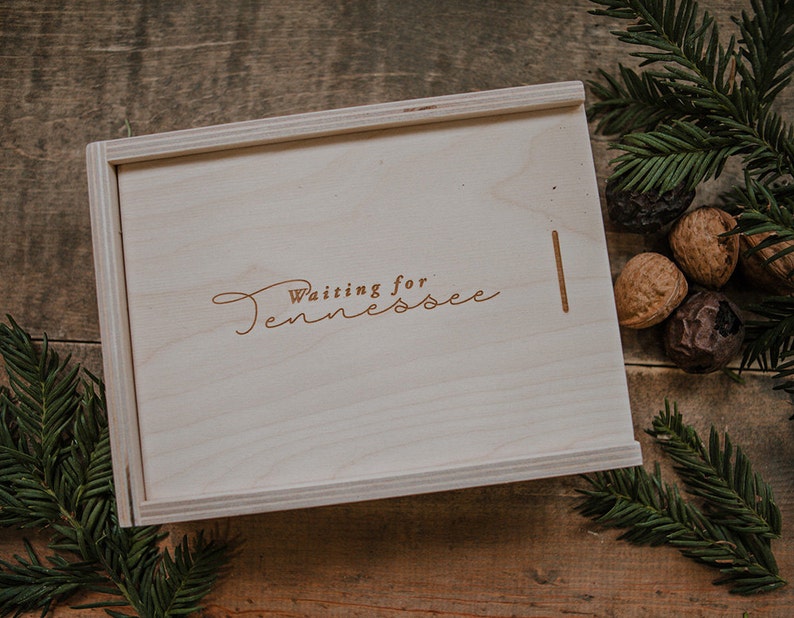 May include: A wooden box with the words "Waiting for Tennessee" engraved on the lid. The box is sitting on a wooden surface with pine branches and walnuts.