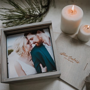 May include: A gray wooden box with a hinged lid, containing a photograph of a couple. The box is engraved with the text "Blue Embers Photography". Two white candles are visible in the background.