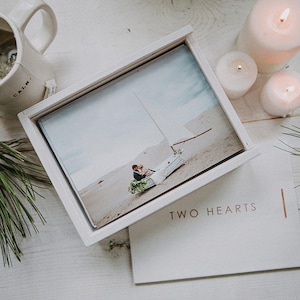 May include: A white wooden photo box with a wedding photo inside, next to a card that reads "TWO HEARTS". A white mug with the word "CALM" is visible, along with lit candles and pine needles, creating a serene, romantic scene.