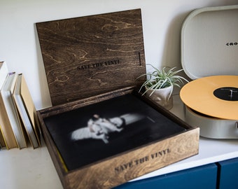 Puede incluir: Una caja de almacenamiento de discos de madera oscura con el texto "SAVE THE VINYL" está abierta, revelando un disco de vinilo. Un disco de vinilo de color dorado se encuentra en un tocadiscos de estilo vintage. También se ve una pequeña planta en una maceta blanca.