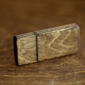Puede incluir: Una peque&ntilde;a caja rectangular de madera con una mancha marr&oacute;n oscura. La caja tiene un patr&oacute;n de veta de madera natural y una fina l&iacute;nea negra que recorre el centro.