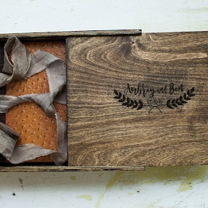 May include: A wooden box with a hinged lid, the lid is open and the inside is lined with brown leather. The lid is engraved with the text "Aubrey and Ben 07.16.16" in a cursive font.