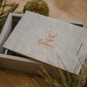 May include: A gray wooden box with a hinged lid. The lid has a logo for "Autumn Woodworks" with a stylized deer design. The box is sitting on a burlap surface.