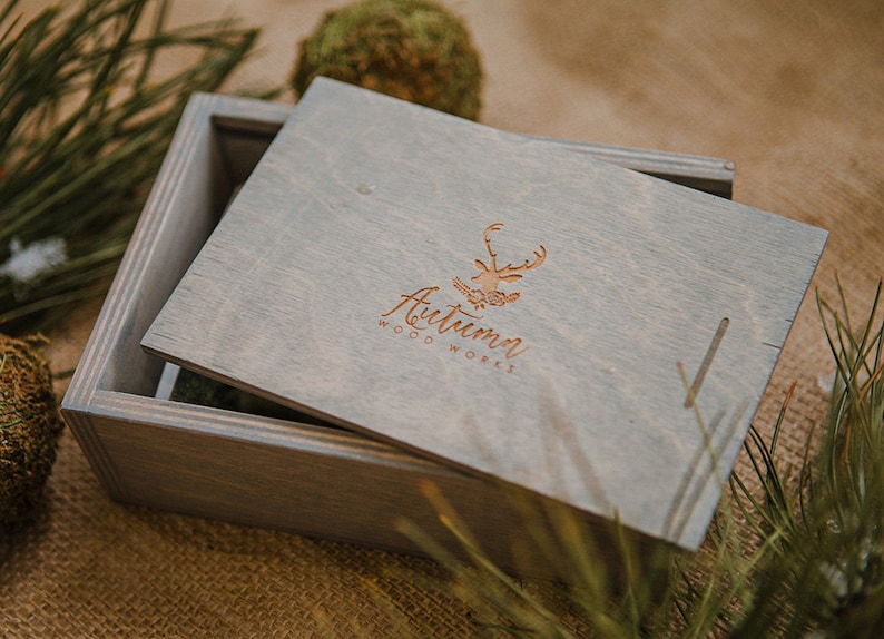 May include: A gray wooden box with a hinged lid. The lid has a logo for "Autumn Woodworks" with a stylized deer design. The box is sitting on a burlap surface.