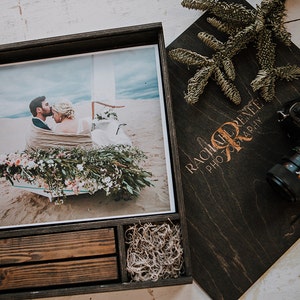 May include: A wooden photo box with a framed wedding photo, a wooden cover with "Rachael Renee Photography" text, and a camera. The photo shows a couple wrapped in a blanket, surrounded by flowers. The box has two small drawers.