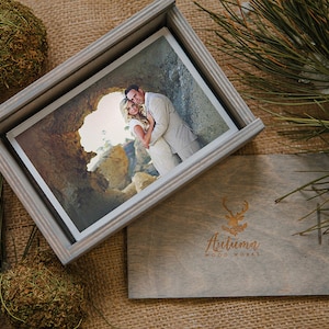 May include: A gray wooden box with a hinged lid, containing a photograph of a couple standing in a rocky landscape. The box has a gold deer logo and the text "Autumn Woodworks" engraved on the front.