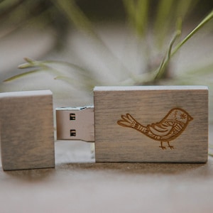 Puede incluir: Una memoria USB de madera con un dise&ntilde;o de p&aacute;jaro grabado en la parte delantera. La memoria est&aacute; cerrada y el puerto USB es visible.