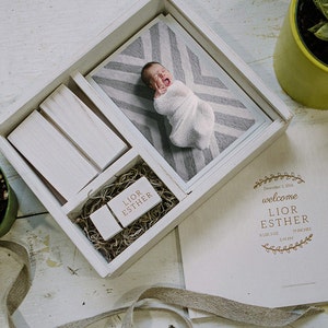 May include: A white wooden box with a gray and white patterned photo of a baby inside. The box also contains a wooden USB drive with the names "Lior Esther" engraved on it. A card with the text "welcome Lior Esther" is on the table next to the box.