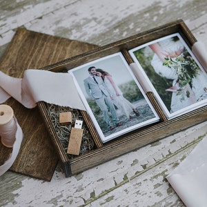 May include: A wooden box with a hinged lid, containing two printed photographs and a wooden USB drive. The box is lined with brown paper and has a light brown ribbon tied around it.