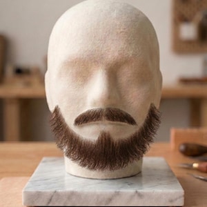 Op de afbeelding: Een beige vilten wollen etalagehoofd met een realistische bruine baard en snor van vezels. Het hoofd is gemonteerd op een vierkante witte marmeren voet met grijze aders. De gelaatstrekken zijn glad en zonder details, met focus op het haar.