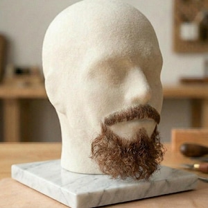 Op de afbeelding: Een gladde, gebroken witte, kale etalagepophoofdsculptuur met een realistische, getextureerde bruine snor en een krullende bruine baard. Het hoofd is gemonteerd op een vierkante, gepolijste witte marmeren voet met subtiele grijze aders in de steen.