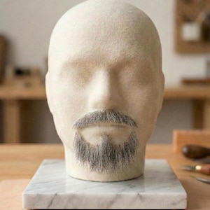 Peut inclure: Une tête de mannequin en laine feutrée beige avec une moustache et un bouc gris réalistes. La tête est montée sur une base carrée en marbre blanc avec des veines grises subtiles. Les traits du visage sont lisses, sans yeux ni oreilles visibles.