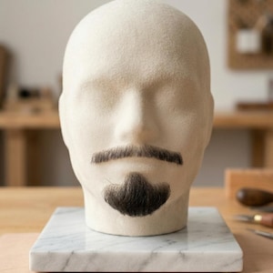 Peut inclure: Une tête de mannequin en feutre beige arborant une moustache brun foncé et un bouc assorti de la même couleur. La tête est fixée sur un socle carré en marbre blanc veiné de gris, offrant une base stable pour présenter ces postiches faciaux.