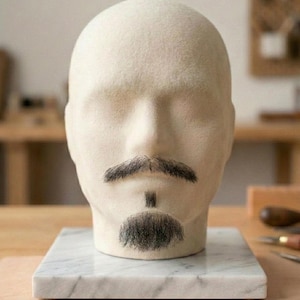 Peut inclure: Une tête de mannequin en laine feutrée beige avec une moustache et un petit bouc brun foncé. La tête est montée sur une base carrée en marbre blanc avec des veines grises. Les traits du visage sont lisses et neutres, avec les poils appliqués.