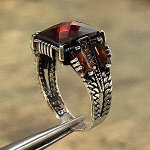 May include: A sterling silver ring featuring a large, faceted, square-cut dark red gemstone at the center, flanked by smaller rectangular red stones. The band displays intricate, textured metalwork and is stamped with the 925 hallmark inside.