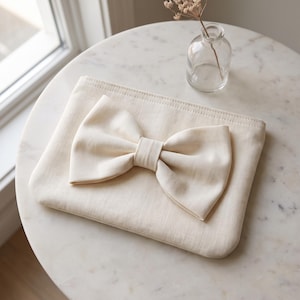 May include: A rectangular off-white fabric pouch featuring a large, centred, matching fabric bow attached to the front. The pouch has a clean, minimalist design with visible stitching along the top edge and a smooth, uniform texture throughout.