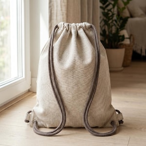 Könnte beinhalten: Ein beigefarbener Kordelzugrucksack aus strukturiertem Stoff. Die Tasche verfügt über einen gerafften Verschluss oben und zwei dicke, dunkelbraune Seilträger, die von den unteren Ecken nach oben verlaufen und ein schlichtes Design bilden.