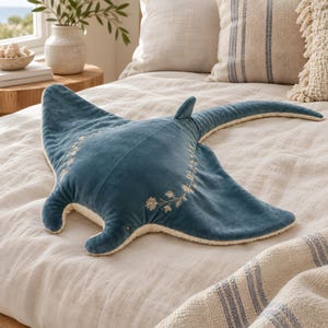 May include: A plush stingray-shaped pillow featuring a soft, deep blue velvet top surface with delicate white floral embroidery along the wings. The underside is crafted from a textured, cream-colored fabric, and the pillow has a long, slender tail.