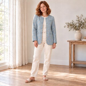 Könnte beinhalten: Ein hellblauer Blazer mit dezentem Blumenmuster, rundem Ausschnitt, langen Ärmeln und einer Knopfleiste auf der Vorderseite. Der Blazer wird mit einer cremefarbenen Hose und einfachen Pantoletten kombiniert, was ein stimmiges Outfit ergibt.