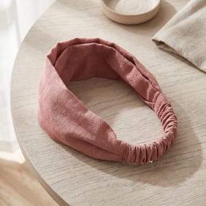 Könnte beinhalten: Ein altrosa Stirnband mit einem breiten, gefalteten Stoffband, das in einen gerafften Gummizug am Hinterkopf übergeht. Das Material weist eine sichtbare, fein strukturierte Webart auf, die dem gesamten Accessoire ein weiches, mattes Aussehen verleiht.