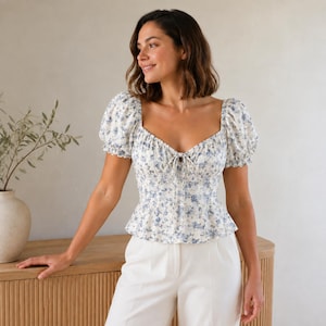May include: A white blouse featuring a blue floral pattern, short puff sleeves, a sweetheart neckline, and a center front tie detail. The garment is paired with white trousers, creating a coordinated outfit with a fitted bodice and peplum hem.
