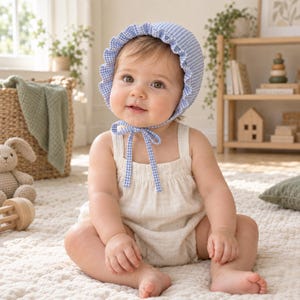 Può includere: Un bambino indossa una cuffietta a quadretti vichy azzurri e bianchi con bordo arricciato e laccetti abbinati, abbinata a una tutina senza maniche color crema. La cuffietta presenta un classico motivo a quadri e si allaccia sotto il mento.