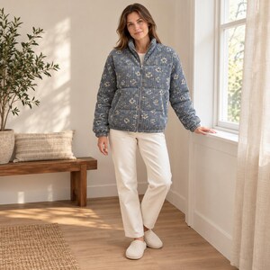 Könnte beinhalten: Eine blaue Steppjacke aus Kord mit weißem Blumenmuster, einem Stehkragen und einem durchgehenden Reißverschluss. Die Jacke wird mit einer schlichten weißen Hose und hellen Slipper-Schuhen kombiniert, was ein abgestimmtes Outfit ergibt.