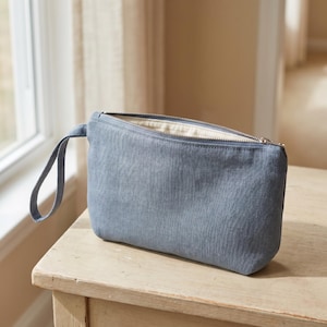 Könnte beinhalten: Eine schieferblaue Reißverschlusstasche mit passender Handschlaufe aus Stoff. Die rechteckige Tasche hat ein strukturiertes Außenmaterial und einen silberfarbenen Metallreißverschluss an der Oberkante, der ein helles Innenfutter zeigt.