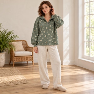 Könnte beinhalten: Ein salbeigrüner Langarm-Pullover mit weißem Gänseblümchenmuster und Viertelreißverschluss, kombiniert mit einer locker sitzenden, hellbeigen melierten Freizeithose und passenden beigen Schlupfschuhen.