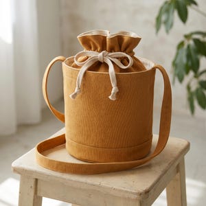 Könnte beinhalten: Eine senfgelbe Beuteltasche mit zylindrischer Form und Kordelzugverschluss. Die Tasche verfügt über eine dicke, cremeweiße gewebte Kordel zum Zuziehen der Öffnung und einen passenden senfgelben Schulterriemen, der an den Seiten befestigt ist.