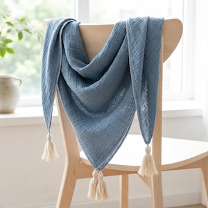 Könnte beinhalten: Ein dreieckiges blaues Musselintuch mit knittriger Textur und drei cremeweißen Quasten an jeder Ecke. Der Stoff ist leicht und halbtransparent und über die Rückenlehne eines hellen Holzstuhls drapiert.