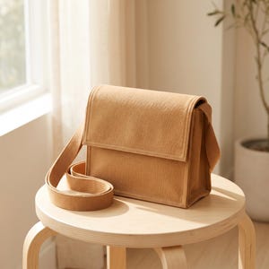 Könnte beinhalten: Eine beigefarbene, rechteckige Umhängetasche mit strukturiertem Klappverschluss und einem passenden gewebten Schulterriemen. Die Tasche hat ein schlichtes, einfarbiges Äußeres mit sichtbaren Nähten an den Kanten und eine kastenförmige, aufrechte Form.