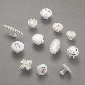May include: A collection of eleven white ceramic cabinet knobs and pulls featuring various designs including gold filigree, floral patterns, metallic rims, and textured edges. The hardware includes round, oval, and fleur-de-lis shapes with metal bases.
