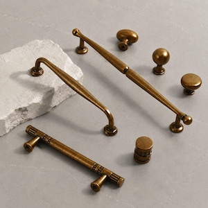 May include: A collection of antique brass cabinet hardware including two long arched handles, one straight handle with ribbed detailing, and three round knobs. The hardware features a weathered metallic finish with visible dark patina accents.