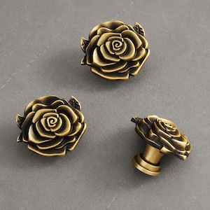 May include: Three drawer knobs shaped like blooming roses with a dark antique brass finish. Each knob features layered, sculpted petals and a central spiral, with a cylindrical base extending from the back of one knob to show the mounting structure.